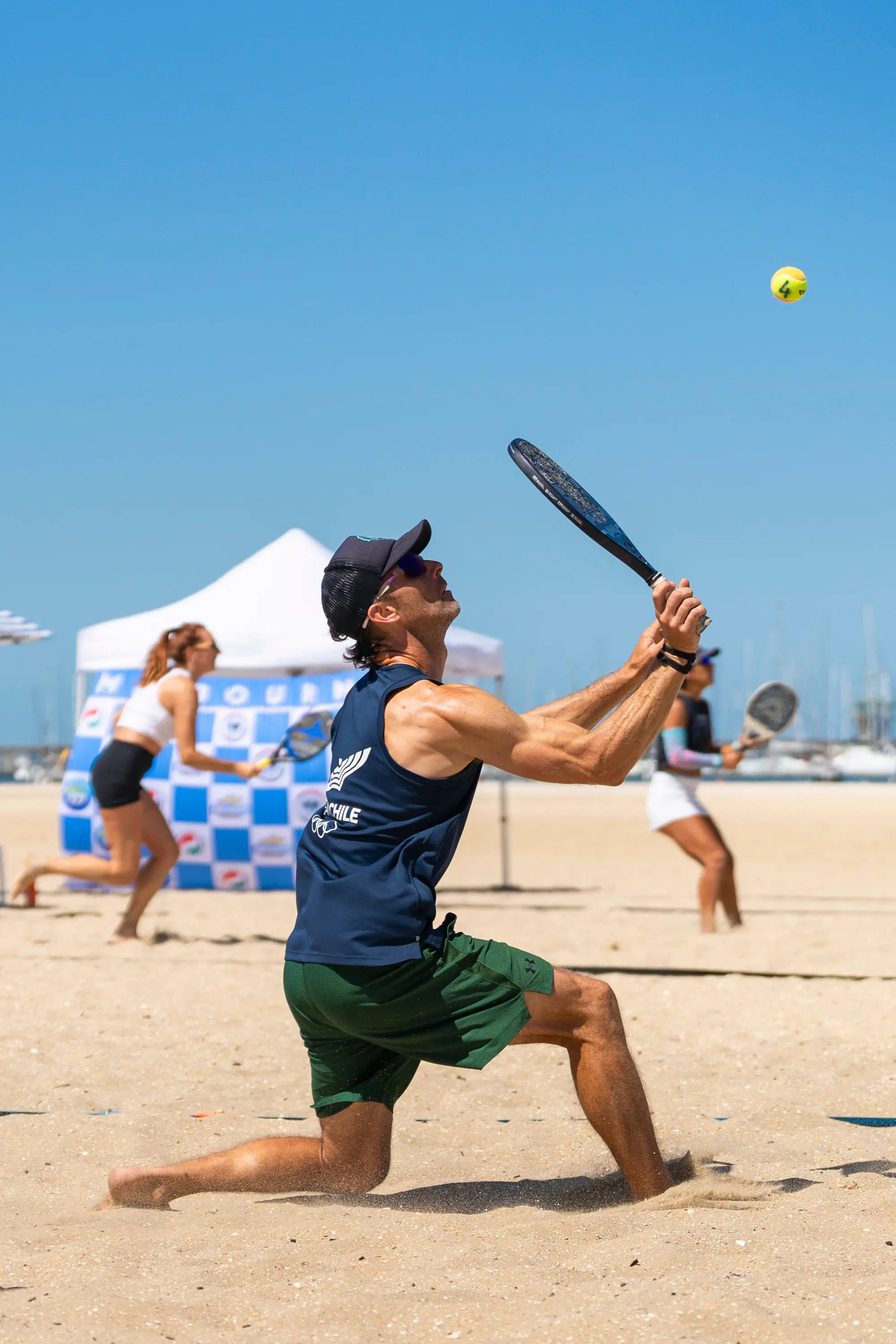 A player lunging low through the sand to reach a ball during a point, with others playing behind