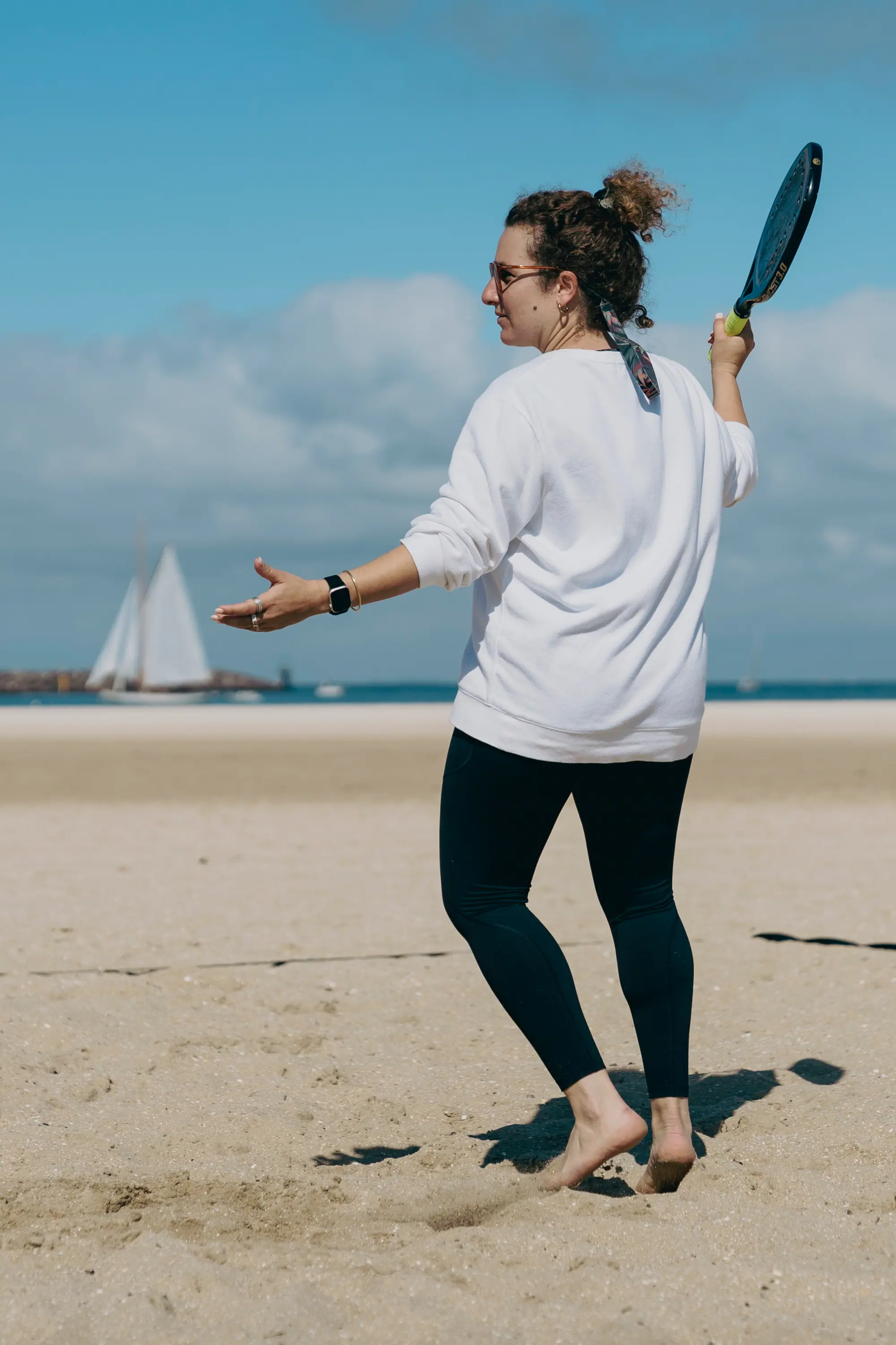 A player mid-swing on the sand, racquet drawn back and free arm pointing at the ball, with the bay behind