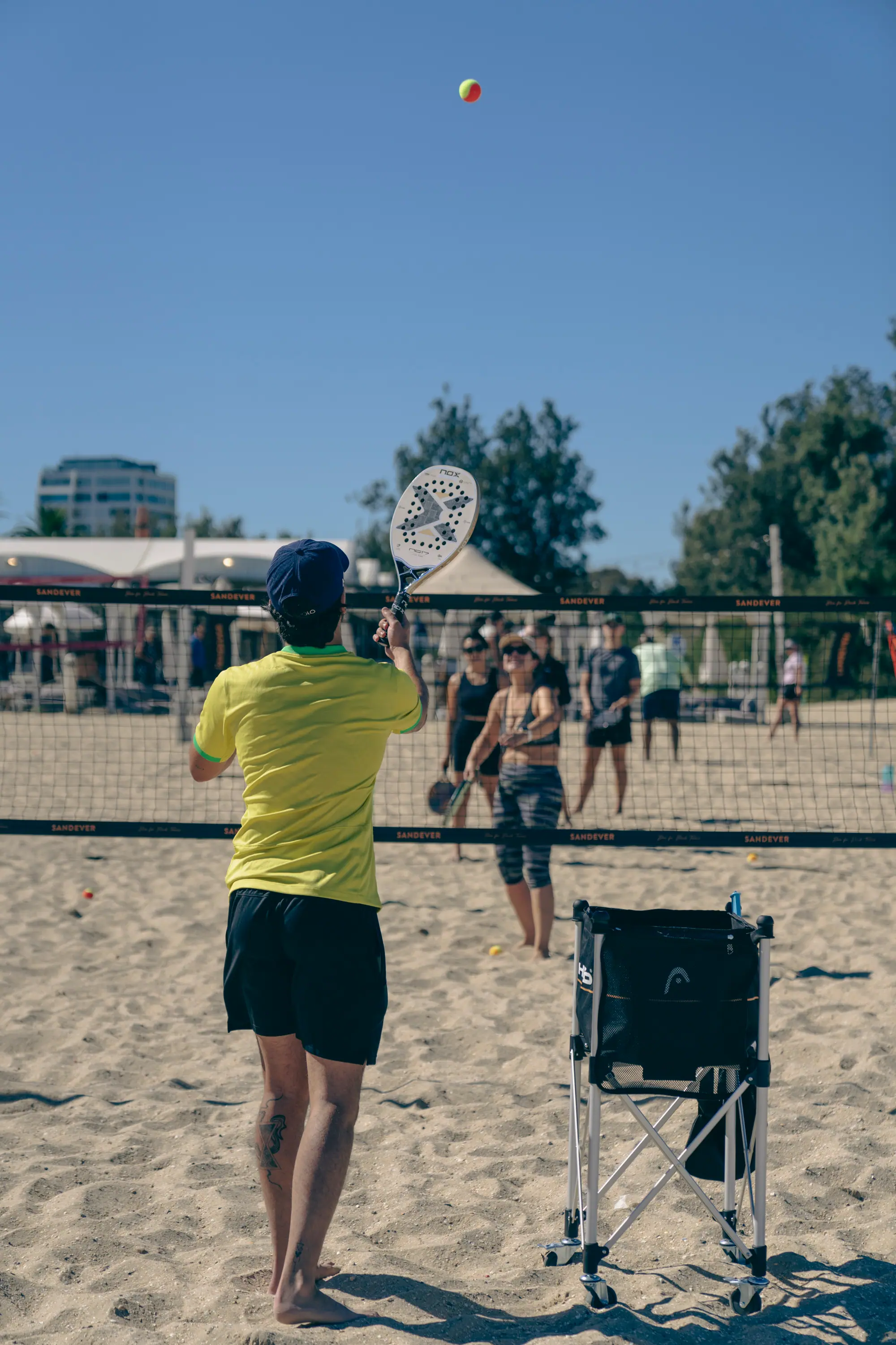 A coach serving a ball over the net to a group waiting on the far side, on the sand at West Beach