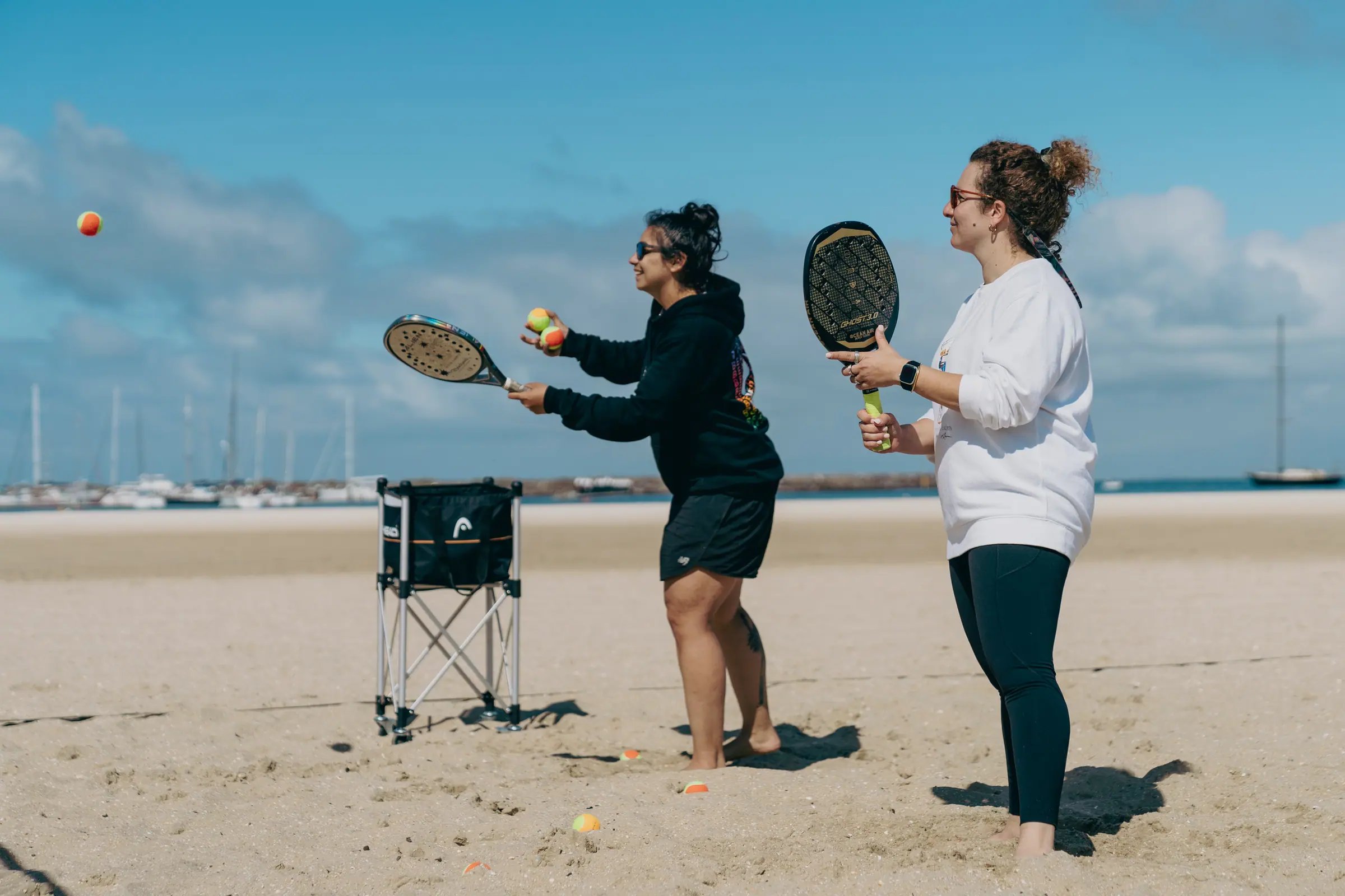 Two players on the sand at West Beach, one feeding a ball to the other, with the St Kilda marina behind them