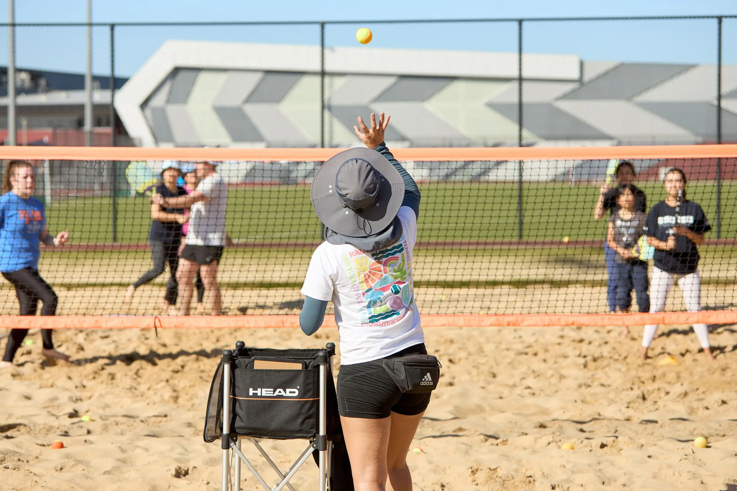 A coach feeding a ball to a group spread along the net on a sand court