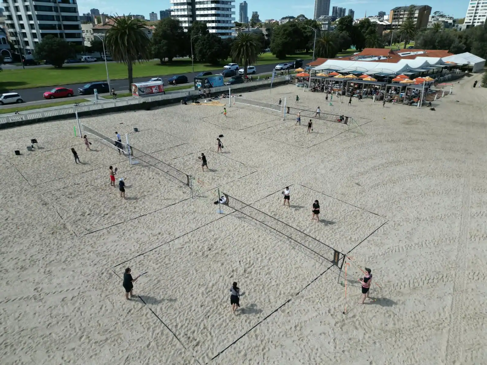 The beach tennis courts on the sand at West Beach, St Kilda, seen from above — several nets in play, with the West Beach Pavilion and the St Kilda skyline behind