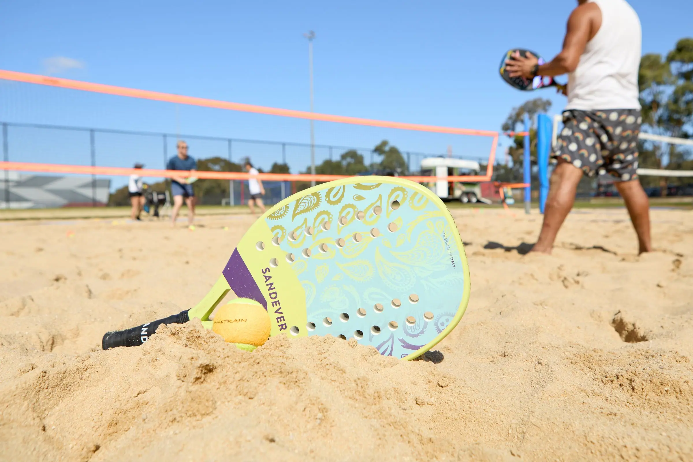 A beach tennis racquet and ball resting in the sand, with players at the net behind