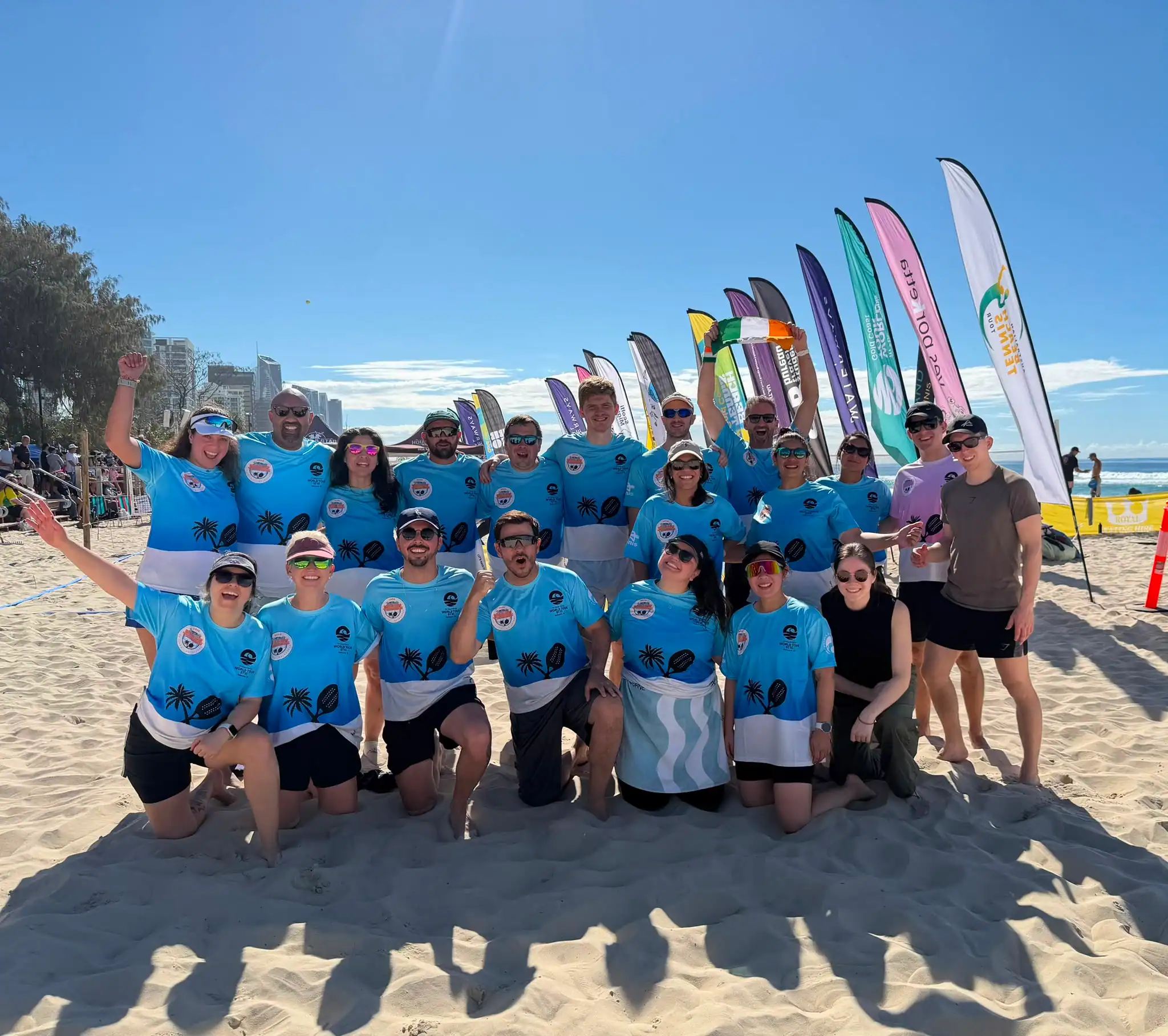 A large group of players in matching club shirts on the sand after a tournament, sponsor flags behind them